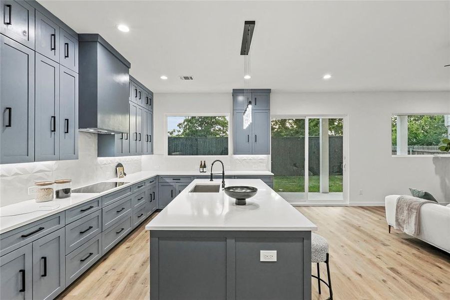 Kitchen featuring wall chimney range hood, light hardwood / wood-style floors, decorative backsplash, black electric cooktop, and an island with sink