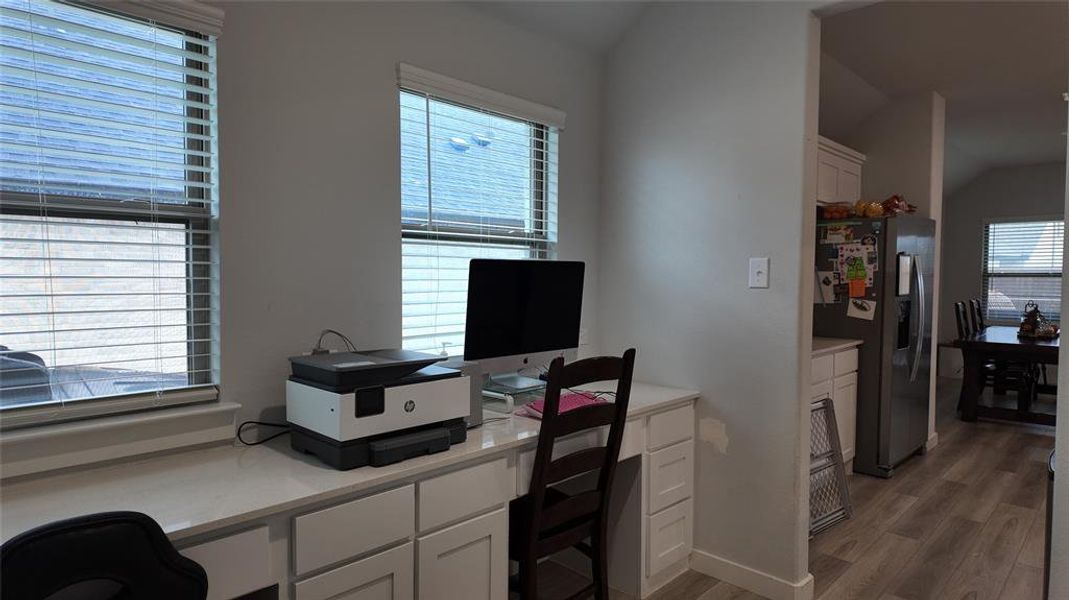 This space features a built-in desk with white cabinetry and a light-colored countertop, illuminated by two windows with blinds