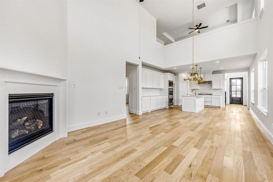 Unfurnished living room featuring a glass covered fireplace, light wood finished floors, a chandelier, ceiling fan, and a high ceiling