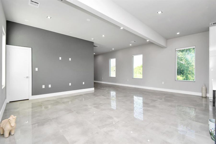 This is a spacious, modern living area with high ceilings, large windows for natural light, and sleek, polished tile flooring. The room features a neutral color palette with a contrast accent wall and is equipped with recessed lighting. There is a staircase in the corner, suggesting a multi-level home.