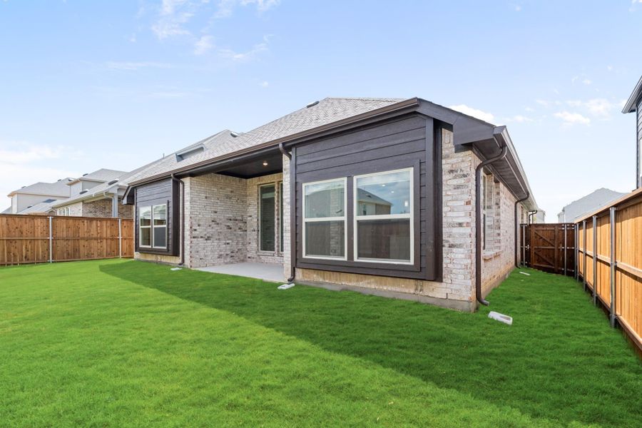 Rear exterior of single-story brick home with dark siding, covered patio, and fenced backyard with green lawn Rear exterior of single-story brick home with dark siding, covered patio, and fenced backyard with green lawn