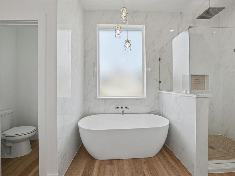 Freestanding soaking tub with wall-mounted faucet, surrounded by marbled-finish tile walls and wood-finish flooring