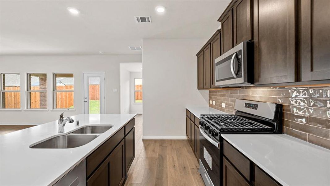 Kitchen featuring wood-finish flooring, dark wood cabinetry, white countertops, stainless steel appliances, and a brown tiled backsplash Kitchen featuring wood-finish flooring, dark wood cabinetry, white countertops, stainless steel appliances, and a brown tiled backsplash