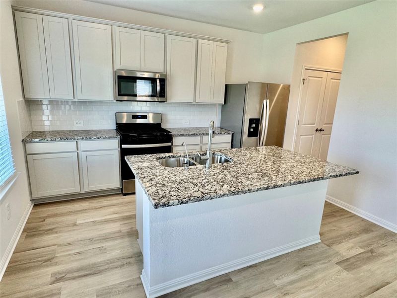 Kitchen featuring stainless steel appliances, light wood-style flooring, a kitchen island with sink, tasteful backsplash, and light stone countertops Kitchen featuring stainless steel appliances, light wood-style flooring, a kitchen island with sink, tasteful backsplash, and light stone countertops