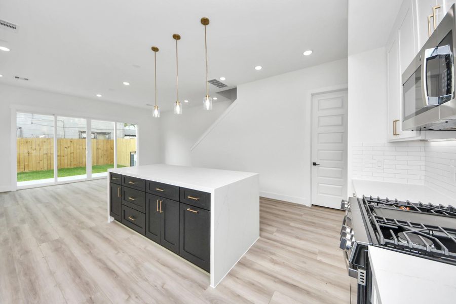 This modern kitchen and living area features an open layout with sleek light fixtures, a large island with dark cabinetry, stainless steel appliances, and natural light from expansive sliding glass doors leading to a fenced backyard.