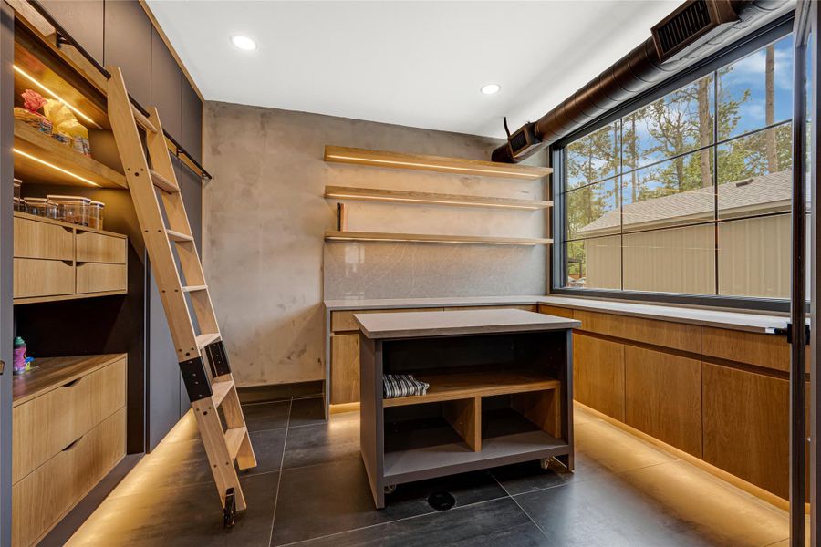 Workspace with open shelving, ample natural light from large windows, and sleek cabinetry. A rolling ladder provides access to high storage, and the neutral tones create a warm, inviting atmosphere.