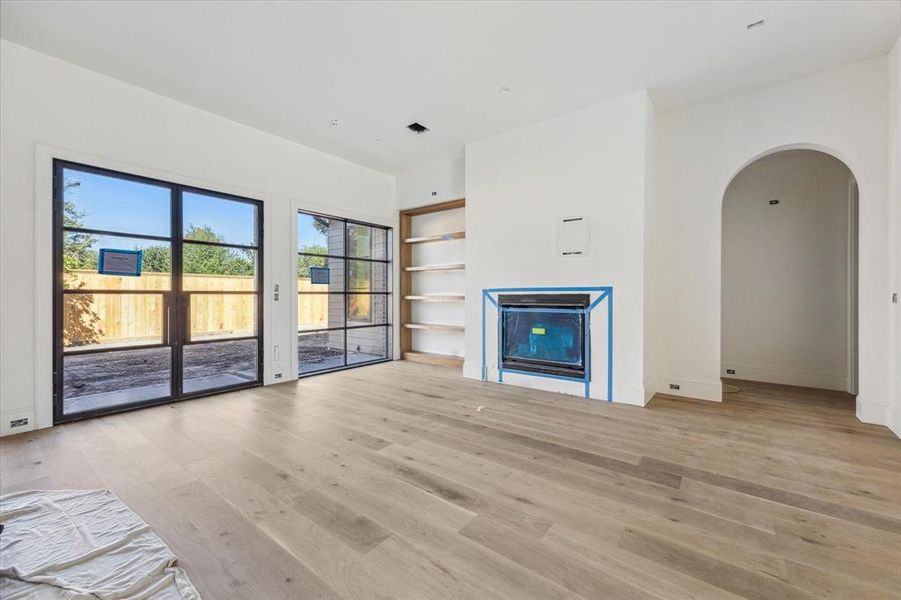 The family room with bright living space and large iron doors leading you to the outdoors, features a gas log fireplace, cast stone mantle, and white oak built-in. The  adjoining archway leads you to the primary suite.