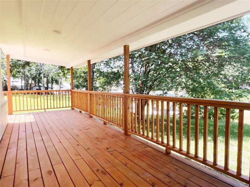 Spacious covered porch with natural wood flooring and railing, featuring a white paneled ceiling and supporting columns