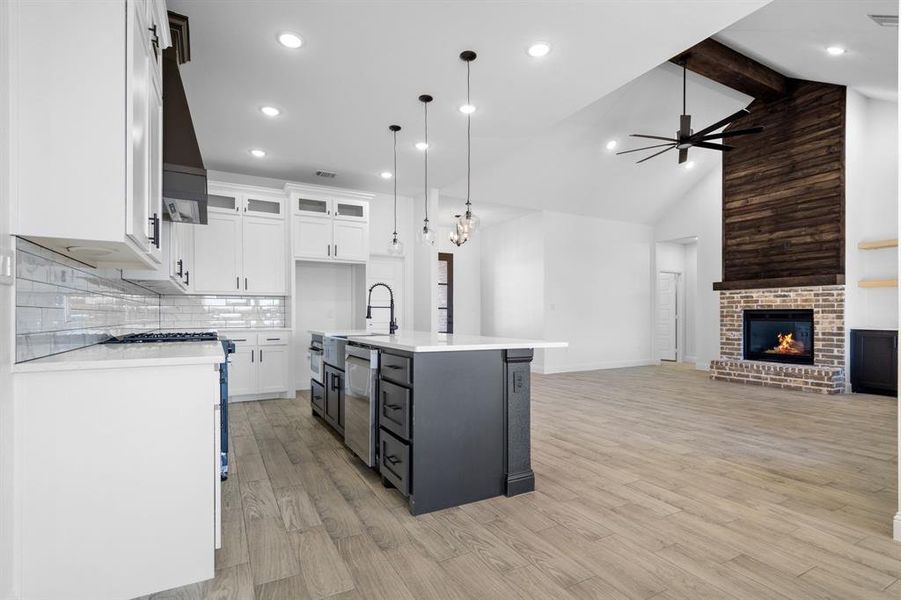 Kitchen featuring decorative light fixtures, a center island with sink, stainless steel dishwasher, a fireplace, and white cabinets