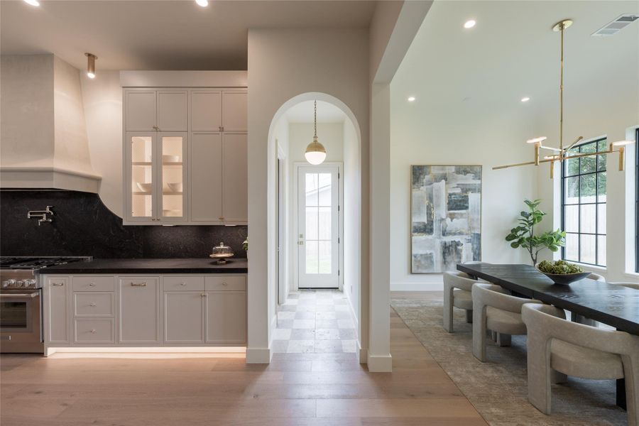 This transitional view captures the artistry of flow—where a softly arched hallway with checkered travertine leads to the backyard, and the dining room opens to the kitchen through a perfectly framed threshold. Each space speaks the same design language, from the curved plaster vent hood and backlit mesh cabinets to the brass fixtures and refined symmetry of the dining area’s barrel-vaulted ceiling. This transitional view captures the artistry of flow—where a softly arched hallway with checkered travertine leads to the backyard, and the dining room opens to the kitchen through a perfectly framed threshold. Each space speaks the same design language, from the curved plaster vent hood and backlit mesh cabinets to the brass fixtures and refined symmetry of the dining area’s barrel-vaulted ceiling.