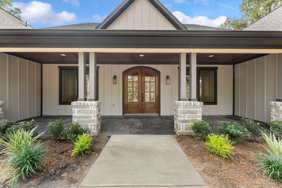 Charming front entry with a covered porch, featuring a wood-paneled ceiling and elegant double doors. The exterior has modern paneling with brick accents, surrounded by landscaping.