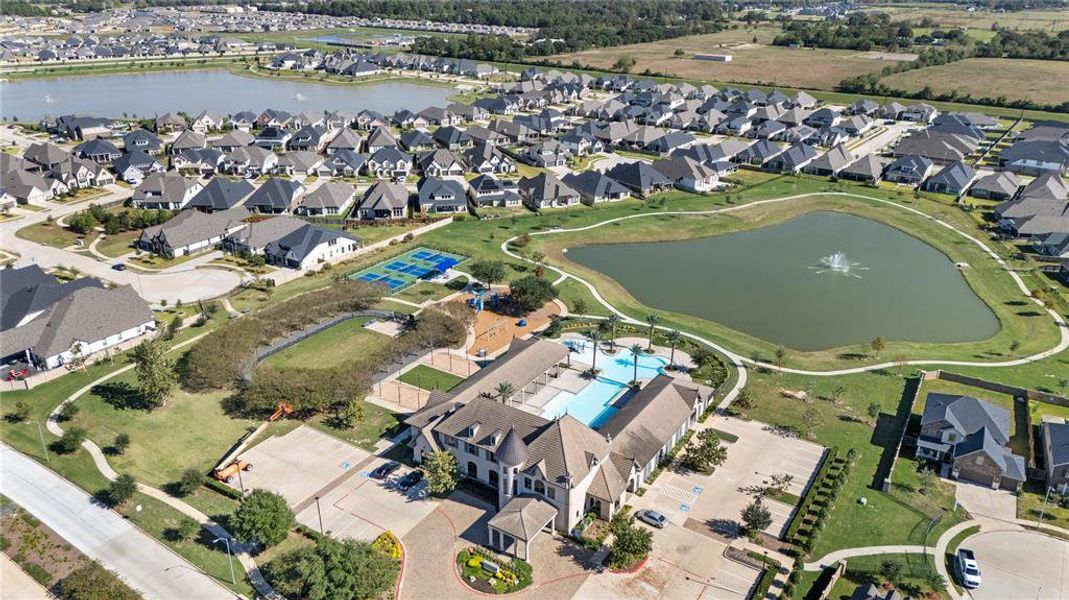 This is an aerial view of a Amira resort community featuring a central pond with a fountain, surrounded by well-spaced single-family homes with gabled roofs. The community includes amenities like a swimming pool and pickleball courts, splash pad (coming soon), fitness center and dog park.