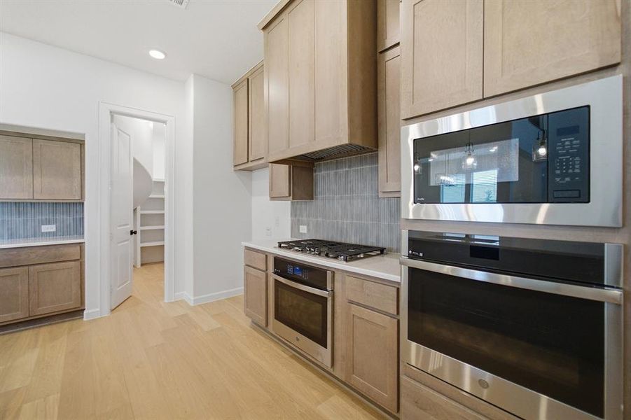 Kitchen featuring light wood-finish cabinetry, stainless steel appliances including a built-in microwave and oven, a gas cooktop, and light countertops with a tile backsplash