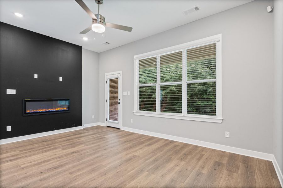 Spacious room featuring wood-finish flooring, a contemporary linear electric fireplace on an accent wall, a ceiling fan with integrated lighting, recessed lighting, and a large window with blinds Spacious room featuring wood-finish flooring, a contemporary linear electric fireplace on an accent wall, a ceiling fan with integrated lighting, recessed lighting, and a large window with blinds