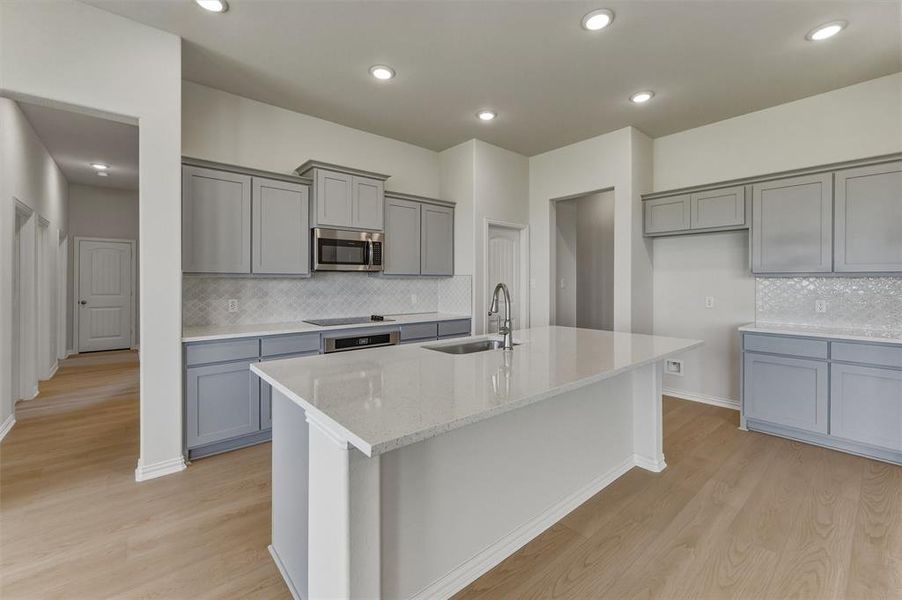 Contemporary kitchen featuring a large central island with a sink, light gray cabinetry, stainless steel microwave, white countertops, and a patterned backsplash