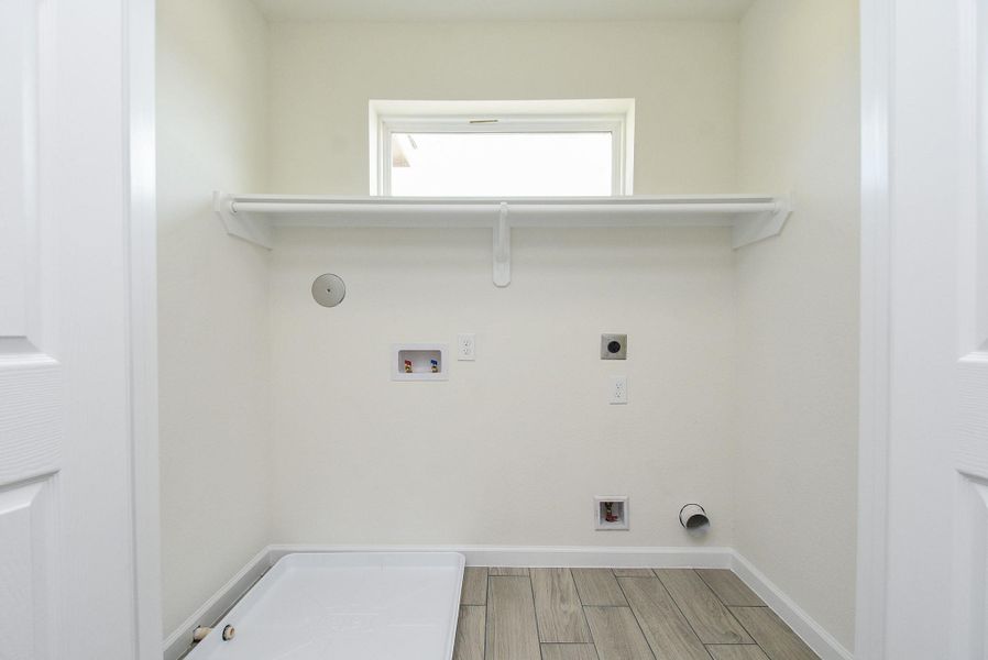 This is a compact laundry room with tile flooring that resembles wood, a small window for natural light, and hookups for a washer and dryer. It includes a shelf for storage above the appliances. This is a compact laundry room with tile flooring that resembles wood, a small window for natural light, and hookups for a washer and dryer. It includes a shelf for storage above the appliances.