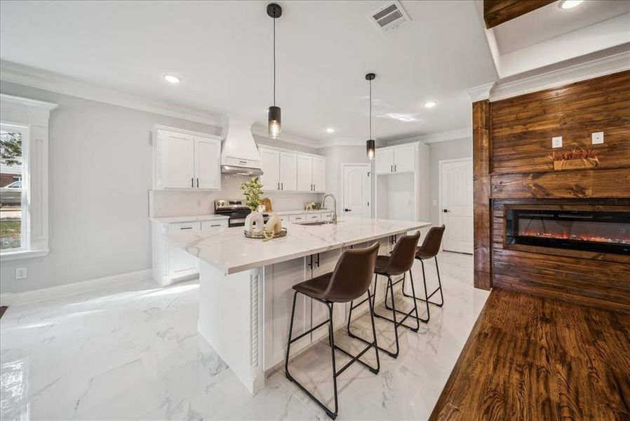 Kitchen featuring stainless steel range with electric stovetop, premium range hood, an island with sink, decorative light fixtures, and white cabinets