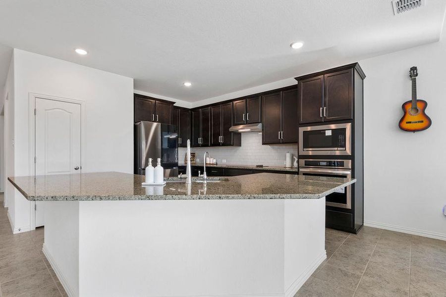 Kitchen with stainless steel appliances, dark stone counters, dark wood finish cabinets, an island with sink, and tasteful backsplash Kitchen with stainless steel appliances, dark stone counters, dark wood finish cabinets, an island with sink, and tasteful backsplash