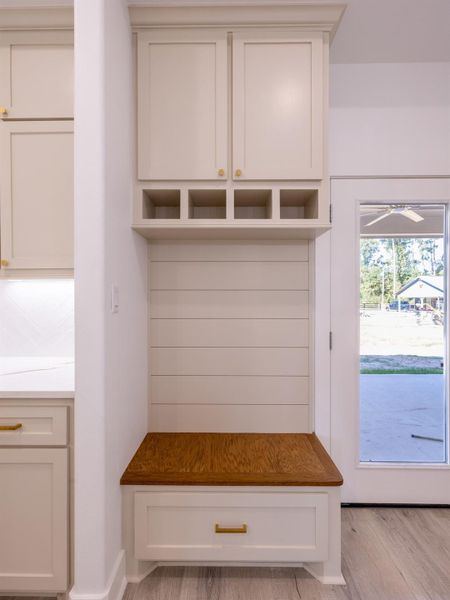 This photo showcases a cozy mudroom corner with custom cabinetry. It features upper cabinets for storage, open cubbies, a wooden bench, and a drawer below for additional storage. The area is bright, with a door leading outside, perfect for organizing shoes and coats.
