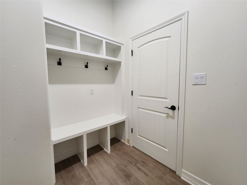 Built-in white storage bench with upper cubbies and matte black hooks, wood-finish flooring, and white paneled door with black hardware