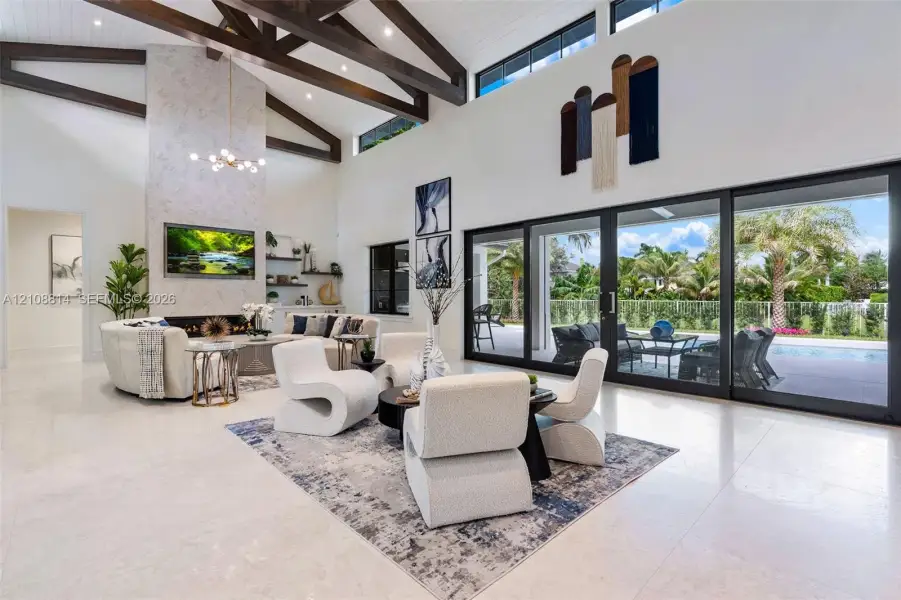 High-ceilinged living area featuring exposed wood ceiling beams, a floor-to-ceiling stone-clad fireplace, built-in shelving, and expansive multi-panel sliding glass doors High-ceilinged living area featuring exposed wood ceiling beams, a floor-to-ceiling stone-clad fireplace, built-in shelving, and expansive multi-panel sliding glass doors