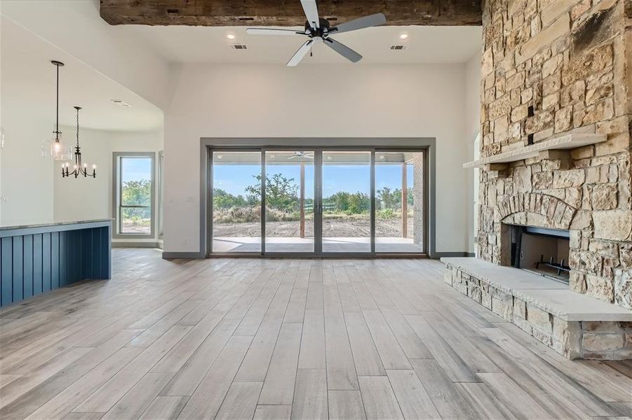 Unfurnished living room featuring light hardwood / wood-style flooring, ceiling fan with notable chandelier, a stone fireplace, beamed ceiling, and a high ceiling