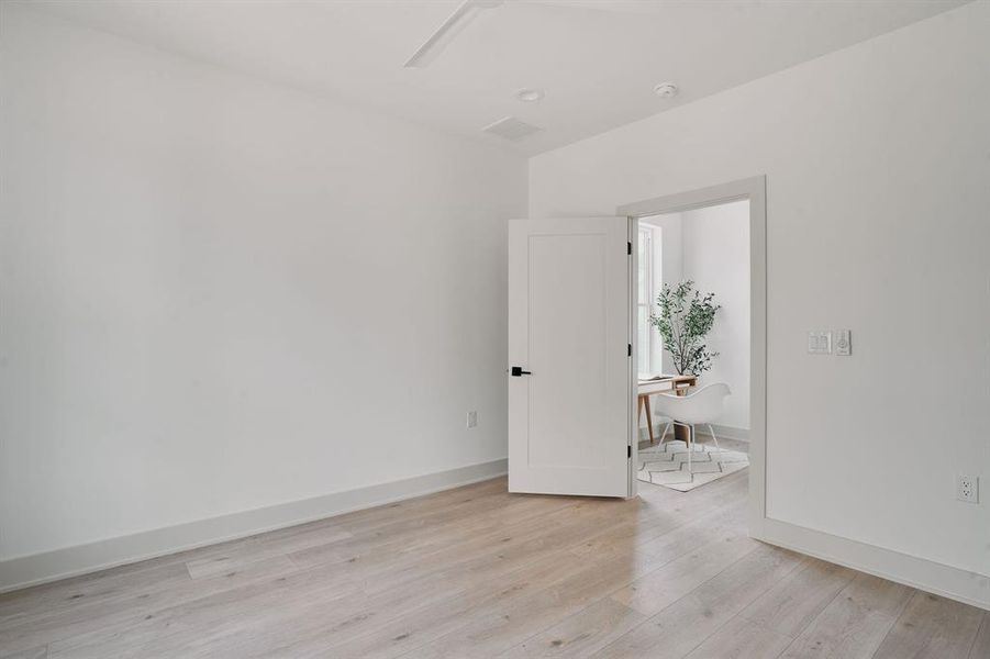 Spacious interior room featuring light wood-finish flooring, white baseboards, and a white interior door with black hardware opening to a separate space
