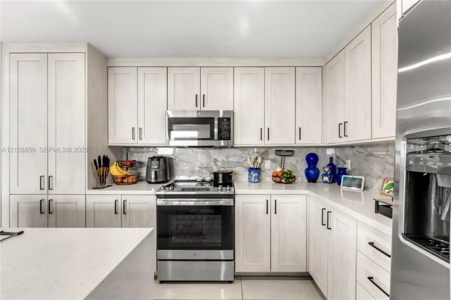 Kitchen featuring light-toned shaker cabinetry, stone-finish backsplash, quartz countertops, stainless steel appliances, and matte black hardware