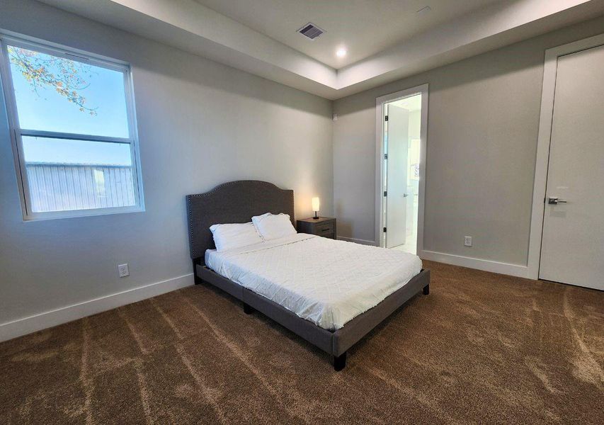 This bedroom features plush carpeting, a modern recessed ceiling with lighting, and a large window providing natural light. It's furnished with a stylish bed and a bedside table with a lamp, and it connects to an adjoining bathroom. This bedroom features plush carpeting, a modern recessed ceiling with lighting, and a large window providing natural light. It's furnished with a stylish bed and a bedside table with a lamp, and it connects to an adjoining bathroom.