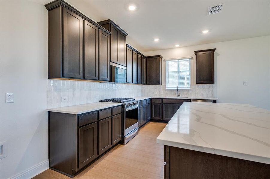 Kitchen with stainless steel gas range, dark brown cabinetry, light stone counters, light wood-type flooring, and decorative backsplash
