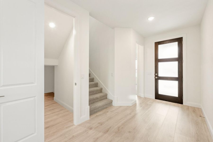 Bright entryway with light wood floors, dark wood front door with glass panels, and carpeted staircase nearby.