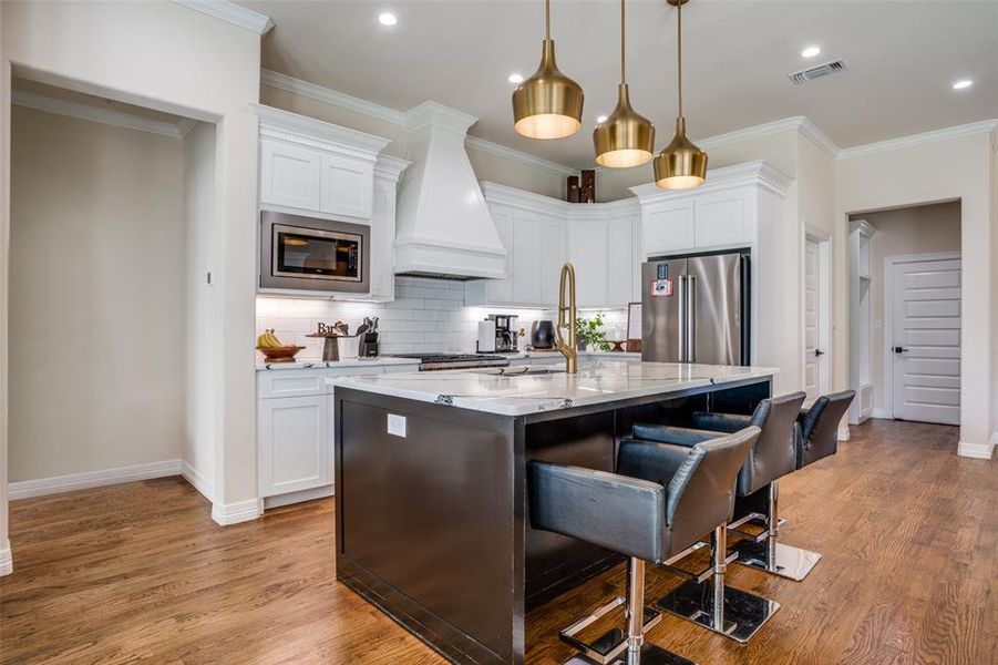 Kitchen featuring appliances with stainless steel finishes, premium range hood, white cabinetry, and a kitchen island with sink