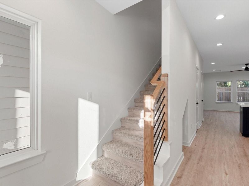 Modern, bright entryway with carpeted stairs, sleek railing, and light wood flooring leading to an open living area with recessed lighting and large windows.