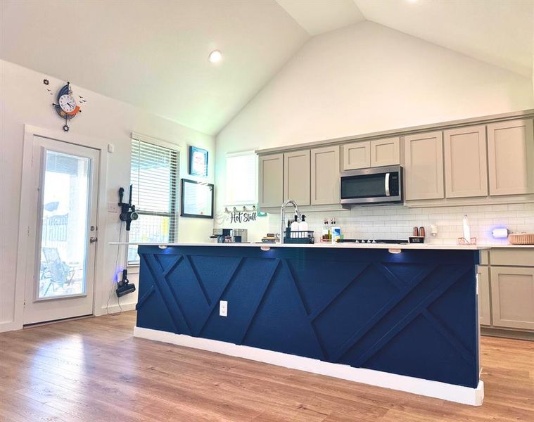 Kitchen featuring gray cabinets, light wood finished floors, backsplash, stainless steel microwave, and high vaulted ceiling