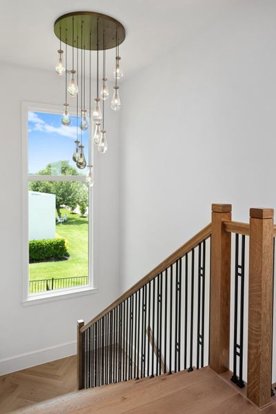 A striking modern chandelier illuminates a well-lit staircase, complemented by views of green landscaping.