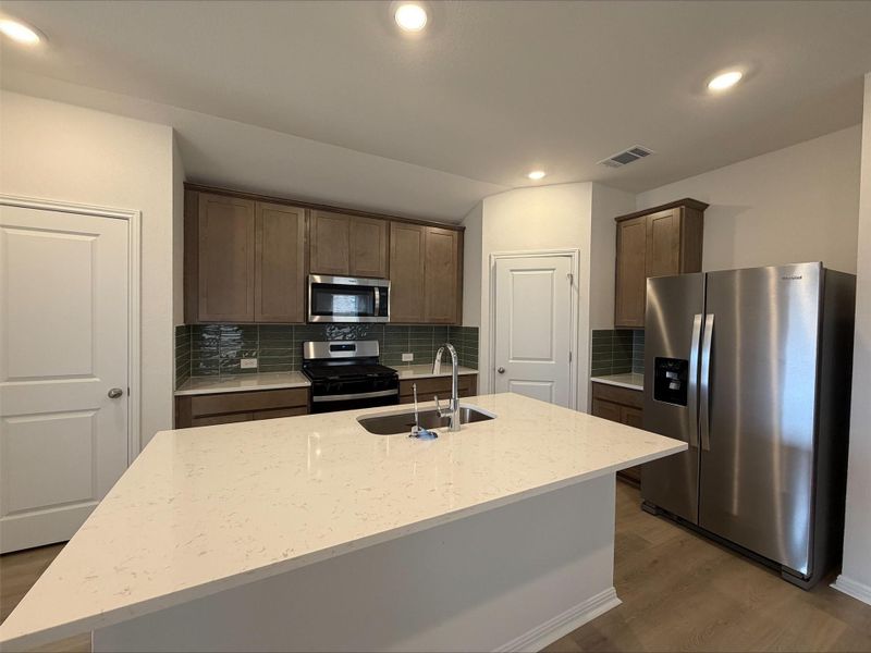 Kitchen with decorative backsplash, appliances with stainless steel finishes, a kitchen island with sink, light stone countertops, and dark wood-style floors