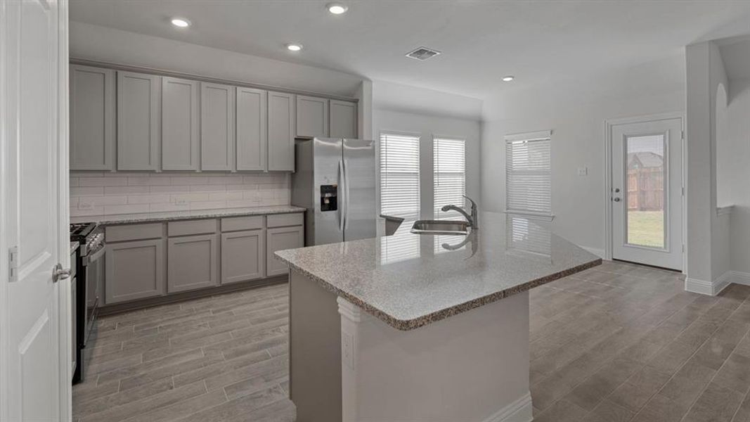 Kitchen with gray cabinetry, tasteful backsplash, stainless steel refrigerator with ice dispenser, light stone countertops, and wood finish floors