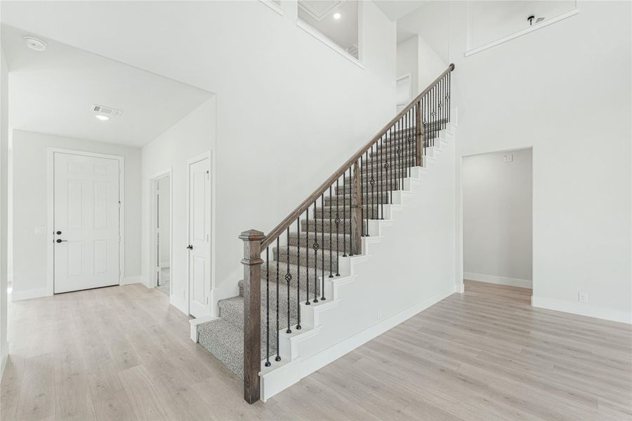 Carpeted staircase with wood handrail and iron balusters in a two-story entryway with light hardwood floors