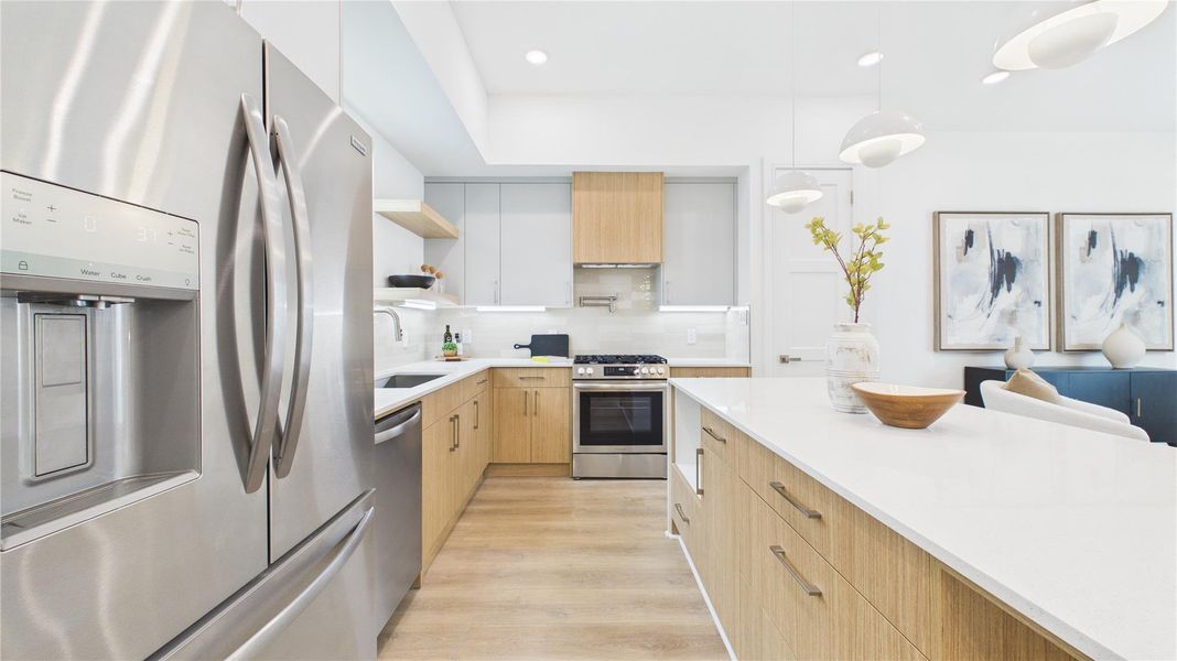 This modern kitchen features sleek stainless steel appliances, light wood cabinetry, and a spacious white countertop island. Pendant lighting and contemporary artwork add elegance, while the open layout enhances functionality and style.