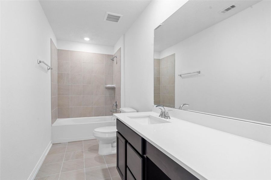 The secondary bathroom showcases tile flooring, a bath/shower combo with Daltile tile surround, a vanity with knee space and stunning light countertop, a mirror, sleek fixtures, and contemporary finishes throughout.