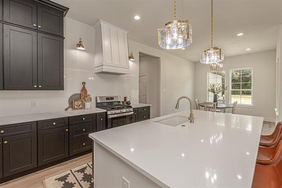 Contemporary kitchen featuring a large center island with an undermount sink, dark cabinetry, light countertops, a stainless steel range, and two decorative pendant light fixtures