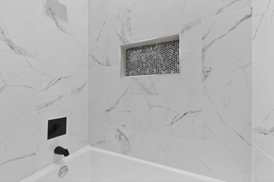 Bathtub with matte black fixtures, surrounded by large format white tile with grey veining, and a recessed niche featuring hexagonal mosaic tile Bathtub with matte black fixtures, surrounded by large format white tile with grey veining, and a recessed niche featuring hexagonal mosaic tile