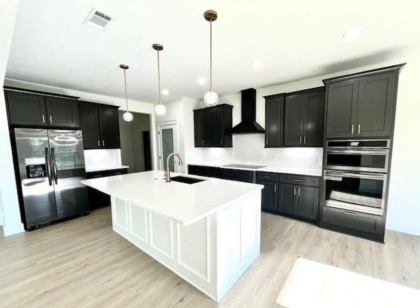 Kitchen with stainless steel appliances, wall chimney exhaust hood, light wood finished floors, a center island with sink, and light countertops