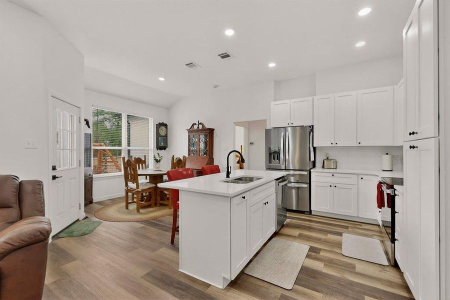 Kitchen featuring a central island with an undermount sink and black faucet, white shaker cabinetry, stainless steel appliances, and wood-finish flooring