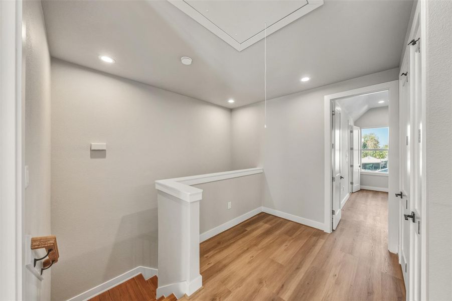 This photo shows a bright upstairs hallway with wood flooring and recessed lighting. It leads to a room with a large window offering a view of the outdoors. The space is modern and inviting, ideal for a cozy home atmosphere.
