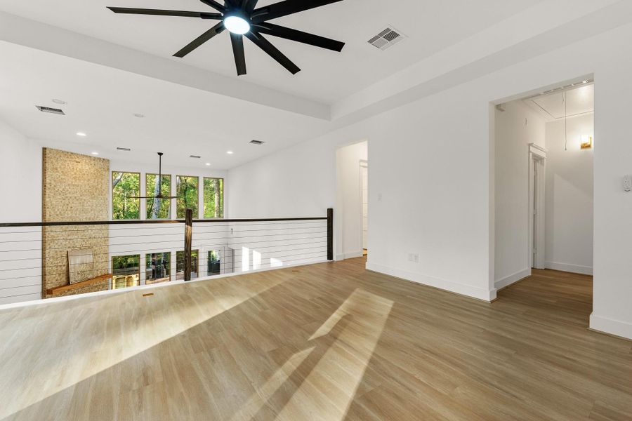 Bright, open loft space with modern design, featuring wood flooring, a ceiling fan, and railing overlooking a spacious living area with large windows and a stone accent wall. Bright, open loft space with modern design, featuring wood flooring, a ceiling fan, and railing overlooking a spacious living area with large windows and a stone accent wall.