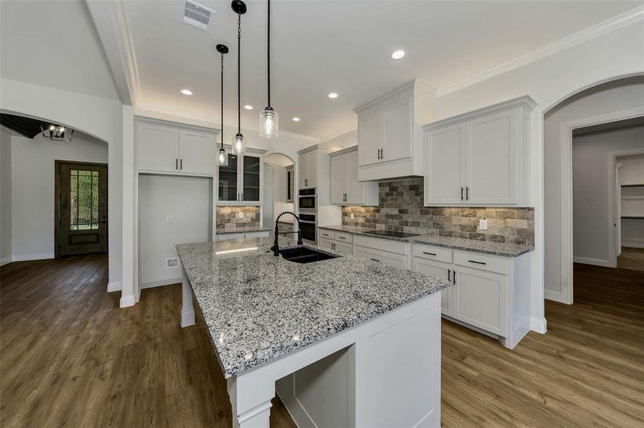Kitchen with crown molding, a kitchen island with sink, arched walkways, light stone counters, and wood finished floors