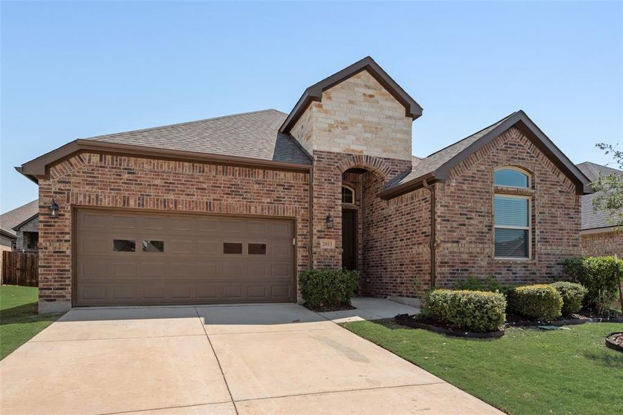 French country inspired facade featuring brick siding, a garage, a shingled roof, concrete driveway, and a front lawn French country inspired facade featuring brick siding, a garage, a shingled roof, concrete driveway, and a front lawn