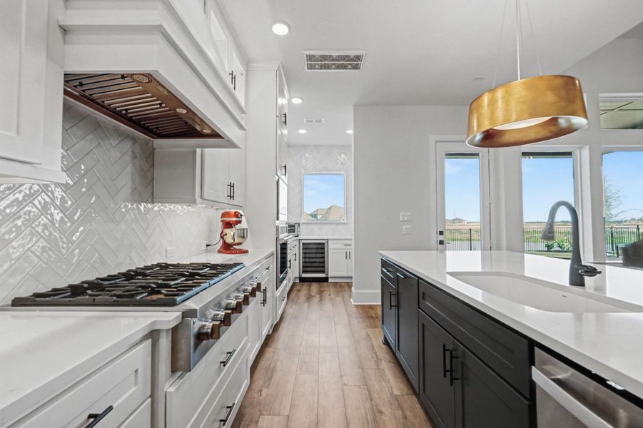 The high-end kitchen is defined by a massive gas cooktop and high-performance range hood. This perspective highlights the custom herringbone backsplash and polished white quartz countertops. Black cabinetry on the island provides a strong visual contrast.