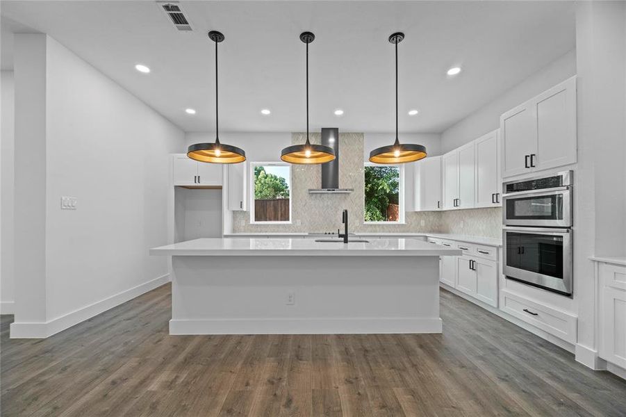 Kitchen featuring stainless steel double oven, wall chimney exhaust hood, tasteful backsplash, light countertops, and recessed lighting Kitchen featuring stainless steel double oven, wall chimney exhaust hood, tasteful backsplash, light countertops, and recessed lighting